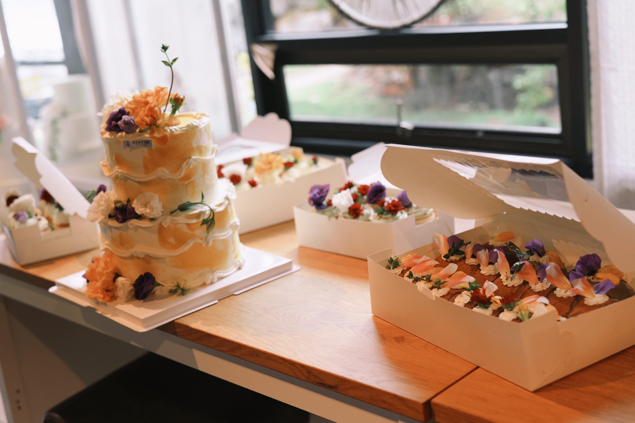A tiered floral wedding cake alongside boxed HoneyMe desserts